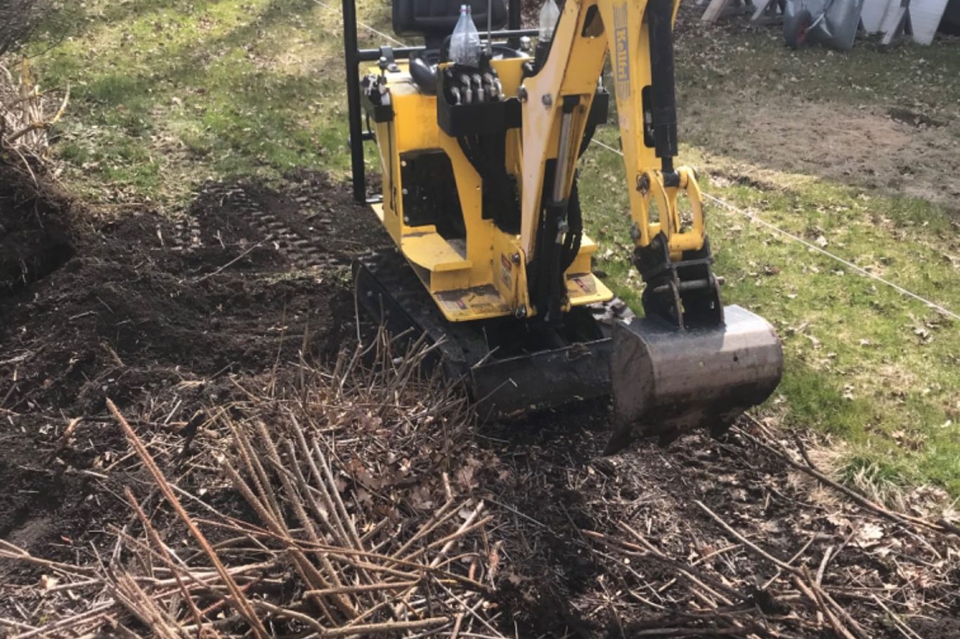 Grävhjälp i trädgårdar Gul grävmaskin arbetar på en jordig yta med strån och grenväxt.