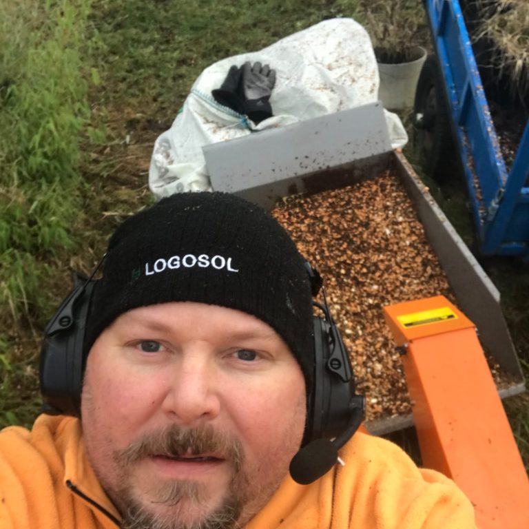 Flisninghjälp Man med hörselskydd tar selfie med en maskin och en hög av spån i bakgrunden.